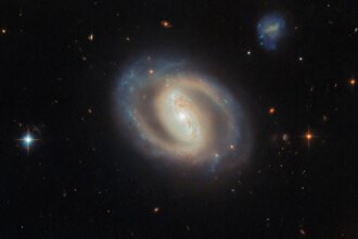 A face-on view of the barred spiral galaxy IC 486, showing a bright, elongated central bar and softly curving, ring-like spiral arms with subtle blue star-forming regions and dark dust lanes, set against a black background dotted with distant galaxies and a few foreground stars.