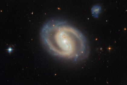 A face-on view of the barred spiral galaxy IC 486, showing a bright, elongated central bar and softly curving, ring-like spiral arms with subtle blue star-forming regions and dark dust lanes, set against a black background dotted with distant galaxies and a few foreground stars.