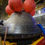 NASA astronaut Christina Koch hugs the Orion spacecraft with her face toward the camera. She is wearing a bright blue jumpsuit. Orion is a rounded cone shaped spacecraft. Its exterior is a sooty black. There are several large orange balloons attached to its top. Orion looks about twice Koch's height.