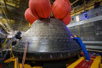 NASA astronaut Christina Koch hugs the Orion spacecraft with her face toward the camera. She is wearing a bright blue jumpsuit. Orion is a rounded cone shaped spacecraft. Its exterior is a sooty black. There are several large orange balloons attached to its top. Orion looks about twice Koch's height.
