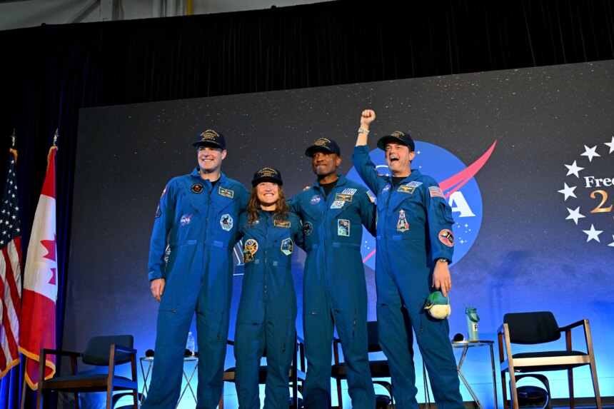 CSA (Canadian Space Agency) astronaut Jeremy Hansen and NASA astronauts Reid Wiseman, Victor Glover, and Christina Koch smile at the crowd during a news conference. They are all wearing blue jumpsuits with patches on the arms and chest areas. Wiseman raises his right fist in a victorious pose.