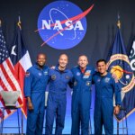 From left to right, NASA astronaut Andre Douglas, ESA (European Space Agency) astronaut Luca Parmitano, and NASA astronauts Randy Bresnik and Frank Rubio take a photo together. Their arms are linked around each other's shoulders. Behind them are the flags of the United States, Texas, NASA, and ESA.