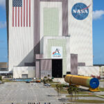 The Artemis III rocket core stage, a massive orange cylinder, moves into the tall Vehicle Assembly Building. A line of people watch from afar on a grassy lawn.