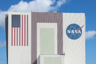 The Artemis III rocket core stage, a massive orange cylinder, moves into the tall Vehicle Assembly Building. A line of people watch from afar on a grassy lawn.