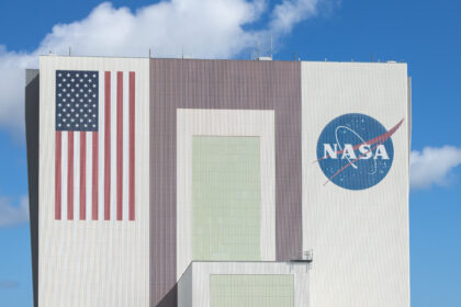 The Artemis III rocket core stage, a massive orange cylinder, moves into the tall Vehicle Assembly Building. A line of people watch from afar on a grassy lawn.