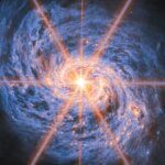 A spiral galaxy shown in mid-infrared light. The image is dominated by an extremely bright glow from the galaxy’s nucleus. Six large and two smaller rays of light emit from the center, which are diffraction spikes created by the telescope’s optics. The galaxy’s spiral arms are visible by two lines of glowing orange bubbles which whirl out into the disc. Swirling blue clouds of dust make up the rest of the galaxy.