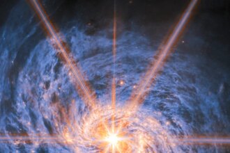 A spiral galaxy shown in mid-infrared light. The image is dominated by an extremely bright glow from the galaxy’s nucleus. Six large and two smaller rays of light emit from the center, which are diffraction spikes created by the telescope’s optics. The galaxy’s spiral arms are visible by two lines of glowing orange bubbles which whirl out into the disc. Swirling blue clouds of dust make up the rest of the galaxy.