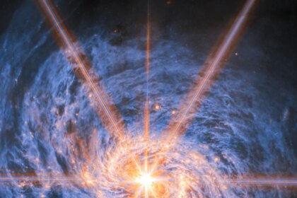 A spiral galaxy shown in mid-infrared light. The image is dominated by an extremely bright glow from the galaxy’s nucleus. Six large and two smaller rays of light emit from the center, which are diffraction spikes created by the telescope’s optics. The galaxy’s spiral arms are visible by two lines of glowing orange bubbles which whirl out into the disc. Swirling blue clouds of dust make up the rest of the galaxy.