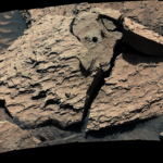 A large, reddish-brown rock as seen by NASA's Curiosity Mars rover. The rock is broken into several pieces. A drill hole is visible in the top middle piece.