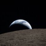 A muted blue Earth with bright white clouds sets behind the cratered lunar surface. The dark portion of Earth is experiencing nighttime. On Earth’s day side, swirling clouds are visible over the Australia and Oceania region. In the foreground, Ohm crater has terraced edges and a flat floor interrupted by central peaks. Central peaks form in complex craters when the lunar surface, liquefied on impact, splashes upwards during the crater’s formation.