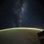 The Milky Way galaxy appears above Earth's greenish atmospheric glow. The galaxy's dust and stars can be seen against the darkness of space.