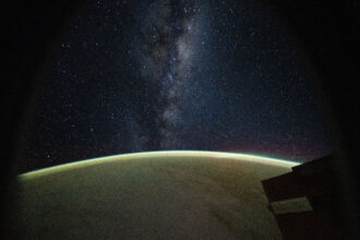 The Milky Way galaxy appears above Earth's greenish atmospheric glow. The galaxy's dust and stars can be seen against the darkness of space.