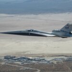 NASA’s X-59 quiet supersonic research aircraft flies above NASA’s Armstrong Flight Research Center in Edwards, California. The plane has a distinctive shape, with a very long, sharp nose. The X-59 is flying fairly low over NASA Armstrong. The compass rose on Rogers Dry Lake at Edwards Air Force Base is visible on the ground beyond NASA Armstrong.