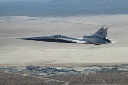 NASA’s X-59 quiet supersonic research aircraft flies above NASA’s Armstrong Flight Research Center in Edwards, California. The plane has a distinctive shape, with a very long, sharp nose. The X-59 is flying fairly low over NASA Armstrong. The compass rose on Rogers Dry Lake at Edwards Air Force Base is visible on the ground beyond NASA Armstrong.