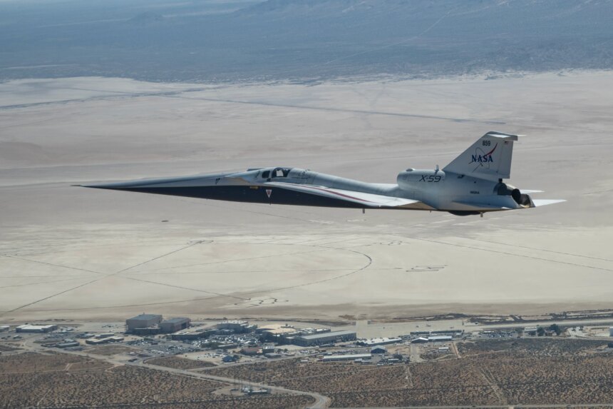 NASA’s X-59 quiet supersonic research aircraft flies above NASA’s Armstrong Flight Research Center in Edwards, California. The plane has a distinctive shape, with a very long, sharp nose. The X-59 is flying fairly low over NASA Armstrong. The compass rose on Rogers Dry Lake at Edwards Air Force Base is visible on the ground beyond NASA Armstrong.