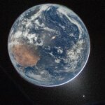 A view of Earth taken from the Orion spacecraft's window. The visible hemisphere appears awash in sunlight, but it is actually lit by moonlight — sunlight reflected from the lunar surface. Along the upper left (south) and lower right (north) edges of Earth’s disk, green auroras glow against the dark sky, showing charged particles from the Sun interacting with gases in Earth’s atmosphere.