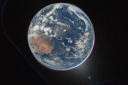 A view of Earth taken from the Orion spacecraft's window. The visible hemisphere appears awash in sunlight, but it is actually lit by moonlight — sunlight reflected from the lunar surface. Along the upper left (south) and lower right (north) edges of Earth’s disk, green auroras glow against the dark sky, showing charged particles from the Sun interacting with gases in Earth’s atmosphere.