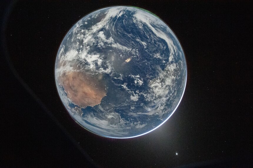 A view of Earth taken from the Orion spacecraft's window. The visible hemisphere appears awash in sunlight, but it is actually lit by moonlight — sunlight reflected from the lunar surface. Along the upper left (south) and lower right (north) edges of Earth’s disk, green auroras glow against the dark sky, showing charged particles from the Sun interacting with gases in Earth’s atmosphere.