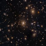 Numerous galaxies dot the scene and appear to cluster around the image center. The view includes large elliptical galaxies along with spiral and lenticular galaxies. Faint arcs of distant galaxies gravitationally lensed by the cluster appear in the upper-right quadrant of the image. A couple of foreground stars are also visible and easily distinguished by their diffraction spikes.