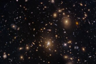 Numerous galaxies dot the scene and appear to cluster around the image center. The view includes large elliptical galaxies along with spiral and lenticular galaxies. Faint arcs of distant galaxies gravitationally lensed by the cluster appear in the upper-right quadrant of the image. A couple of foreground stars are also visible and easily distinguished by their diffraction spikes.