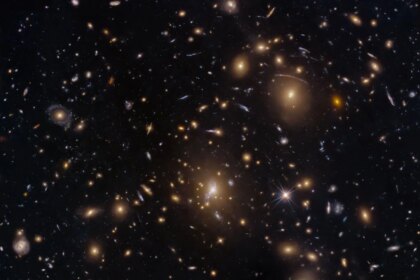 Numerous galaxies dot the scene and appear to cluster around the image center. The view includes large elliptical galaxies along with spiral and lenticular galaxies. Faint arcs of distant galaxies gravitationally lensed by the cluster appear in the upper-right quadrant of the image. A couple of foreground stars are also visible and easily distinguished by their diffraction spikes.