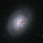 This Hubble image is of a bright lenticular galaxy (NGC 1266) seen nearly face on. Broad bright and dimmer areas of light hint at spiral structure, but there are no distinct spiral arms. Rusty-reddish-brown dust bisects the galaxy. More distant galaxies dot the black background, some even shining through the less dense regions of the bright foreground lenticular galaxy.