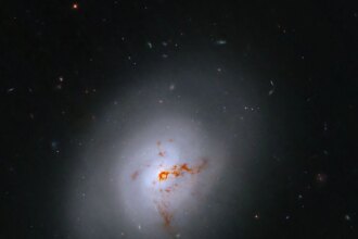 This Hubble image is of a bright lenticular galaxy (NGC 1266) seen nearly face on. Broad bright and dimmer areas of light hint at spiral structure, but there are no distinct spiral arms. Rusty-reddish-brown dust bisects the galaxy. More distant galaxies dot the black background, some even shining through the less dense regions of the bright foreground lenticular galaxy.