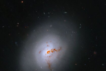 This Hubble image is of a bright lenticular galaxy (NGC 1266) seen nearly face on. Broad bright and dimmer areas of light hint at spiral structure, but there are no distinct spiral arms. Rusty-reddish-brown dust bisects the galaxy. More distant galaxies dot the black background, some even shining through the less dense regions of the bright foreground lenticular galaxy.