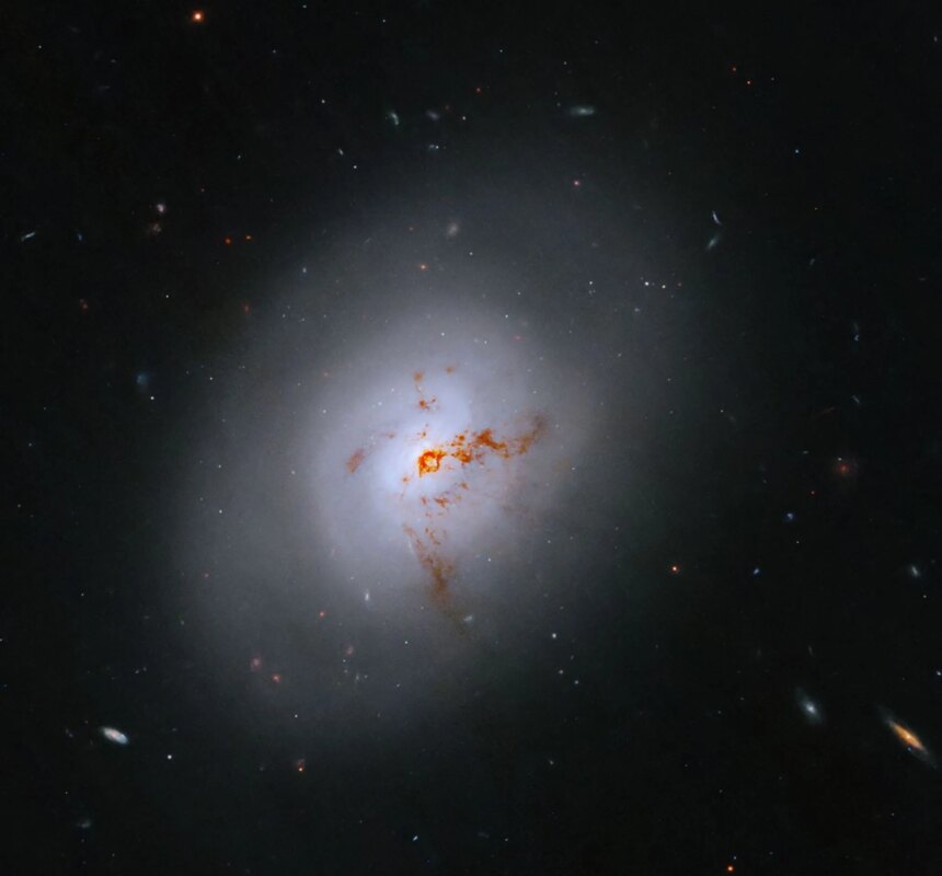 This Hubble image is of a bright lenticular galaxy (NGC 1266) seen nearly face on. Broad bright and dimmer areas of light hint at spiral structure, but there are no distinct spiral arms. Rusty-reddish-brown dust bisects the galaxy. More distant galaxies dot the black background, some even shining through the less dense regions of the bright foreground lenticular galaxy.