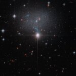 A star in the foreground shines brightly with four diffraction spikes. Above this star is a diffuse and faint dwarf irregular galaxy. The galaxy itself has bright blue and red stars.
