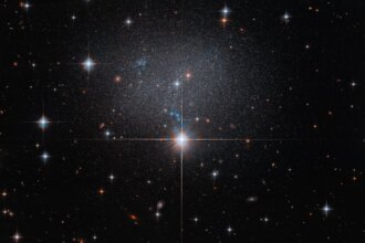 A star in the foreground shines brightly with four diffraction spikes. Above this star is a diffuse and faint dwarf irregular galaxy. The galaxy itself has bright blue and red stars.