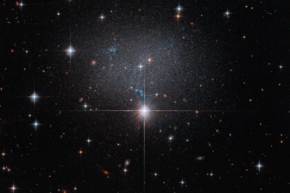 A star in the foreground shines brightly with four diffraction spikes. Above this star is a diffuse and faint dwarf irregular galaxy. The galaxy itself has bright blue and red stars.