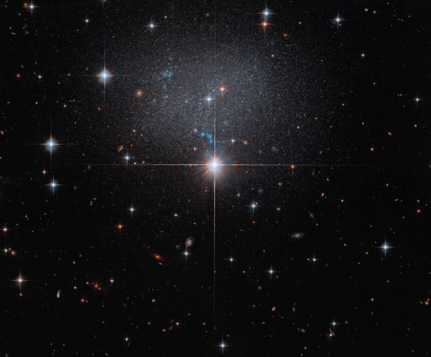 A star in the foreground shines brightly with four diffraction spikes. Above this star is a diffuse and faint dwarf irregular galaxy. The galaxy itself has bright blue and red stars.