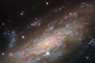 A close view of spiral galaxy that is tilted at an angle, so that its disk fills the view from corner to corner. Its disk is yellow near to the center and pale blue farther out, showing cooler and hotter stars, respectively. Thin brown clouds of dust, glowing pink spots of star formation, and sparkling blue patches filled with star clusters swirl through the galaxy. Behind it, small orange dots are very distant galaxies.