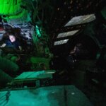 Astronaut Christina Koch is nestled into a corner of the Orion crew capsule, reading. The rest of the capsule is dark, but her face is lit by the glow from her tablet. Two of her other crew members can dimly be seen looking out the window and floating in the foreground.