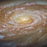 A large spiral galaxy. It is seen tilted at an angle, so that it is foreshortened and appears very wide. Its tightly-wound, blue spiral arms swirl out from its glowing center, spreading apart at the tips. They are followed by strands and clumps of dark red dust and spotted with pink dots where stars are forming in clouds of gas. The galaxy is surrounded by a slight glow and lies on a dark background.