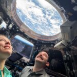Two astronauts look up through a round window in the International Space Station's cupola. Earth's blue water and white clouds are visible. In a trapezoid-shaped window between the two astronauts, we can see the blue haze of Earth's atmosphere.