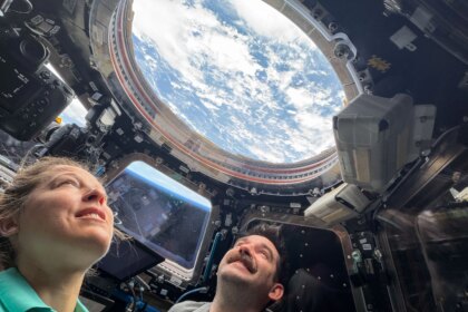 Two astronauts look up through a round window in the International Space Station's cupola. Earth's blue water and white clouds are visible. In a trapezoid-shaped window between the two astronauts, we can see the blue haze of Earth's atmosphere.