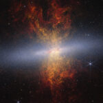 Hourglass-shaped plumes of gas shoot outward from above and below a bright disk-shaped center. The plumes are yellow near the galaxy’s bright center and gradually become redder as you move farther away. M82 is set against the black background of space, which has many distant galaxies that appear as small white and orange spirals, ovals, and points of light. On the right of M82 is a blue-white star with diffraction spikes.
