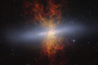 Hourglass-shaped plumes of gas shoot outward from above and below a bright disk-shaped center. The plumes are yellow near the galaxy’s bright center and gradually become redder as you move farther away. M82 is set against the black background of space, which has many distant galaxies that appear as small white and orange spirals, ovals, and points of light. On the right of M82 is a blue-white star with diffraction spikes.