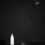 The Moon (right) and Venus (the small dot of light at center) shine above the lit-up Washington Monument at night. Only a small sliver of the Moon is visible.