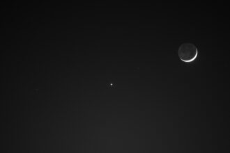 The Moon (right) and Venus (the small dot of light at center) shine above the lit-up Washington Monument at night. Only a small sliver of the Moon is visible.