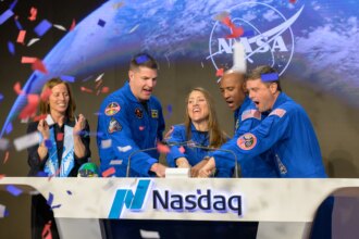 Nasdaq Chair and Chief Executive Officer Adena T. Friedman, left, and NASA’s Artemis II crewmembers CSA (Canadian Space Agency) astronaut Jeremy Hansen, and NASA astronauts Christina Koch, Victor Glover, and Reid Wiseman, right, ring the closing bell of the Nasdaq market session. They're standing behind a wide white podium with the Nasdaq logo and the word "Nasdaq." There is a screen on the front of the podium displaying the NASA meatball logo. Friedman is standing a little further away from the podium. Red, white, and blue confetti falls from the air on the left side of the image.