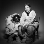 NASA Astronaut Jessica Meir sits in a chair. Her body is slightly turned to the right as she looks forward. She is wearing the bottom half of a spacesuit; the top half and gloves are on the floor next to the chair.