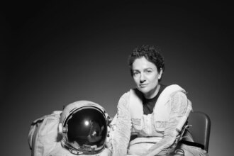 NASA Astronaut Jessica Meir sits in a chair. Her body is slightly turned to the right as she looks forward. She is wearing the bottom half of a spacesuit; the top half and gloves are on the floor next to the chair.