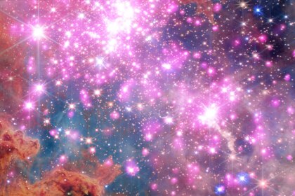 Scores of gleaming stars ringed in neon pink stretch across the frame, highlighting a cluster where stars are between one and three million years old. Brick-orange dust clouds along the bottom edge illustrate the raw materials of this active stellar nursery.
