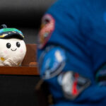 Rise, the Artemis II zero gravity indicator, sits on a wooden desk. A NASA astronaut is in the foreground.