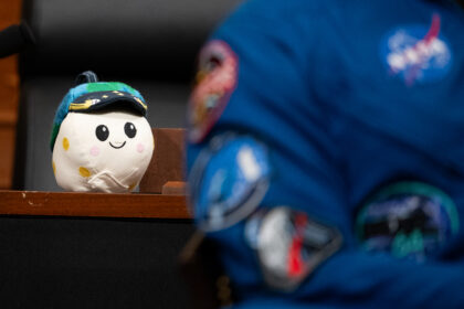 Rise, the Artemis II zero gravity indicator, sits on a wooden desk. A NASA astronaut is in the foreground.