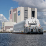 A large flat ship with a rounded structure on it approaches Kennedy Space Center's Vehicle Assembly Building. Dark blue water is in the foreground, while the ship moves toward the building in the background. The sky is blue, but huge, white fluffy clouds covering a good portion of it.