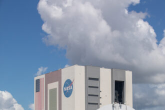 A large flat ship with a rounded structure on it approaches Kennedy Space Center's Vehicle Assembly Building. Dark blue water is in the foreground, while the ship moves toward the building in the background. The sky is blue, but huge, white fluffy clouds covering a good portion of it.